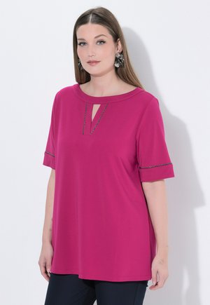 Kvinde med langt brunt hår iført store blomsterøreringe og en magenta top med korte ærmer og nøglehul ved halsen samt sorte bukser.