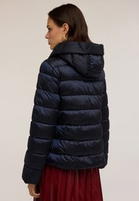 Jachetă puffer bleumarin cu glugă, cu secțiuni orizontale matlasate și o textură netedă, lucioasă. Accents cu manșete ribbed.