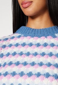 Mujer que lleva un suéter de punto con rayas en forma de concha en azul, rosa, blanco y azul claro, y un cuello acanalado azul.