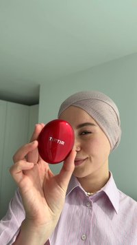 Conteneur cosmétique ovale rouge étiqueté "TIRTIR" tenu dans une main. Surface lisse, finition brillante, design compact. Le fond est d'un vert doux.