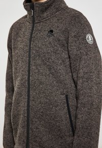 Giacca in pile grigia scura con colletto alto, chiusura con zip, tasche laterali e una patch con logo sulla manica. Presenta un tessuto texturizzato.