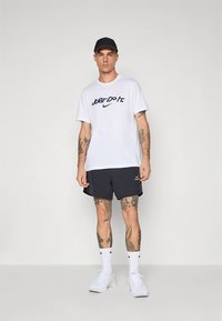 T-shirt branca de algodão com logótipo preto "Just Do It", combinada com calções azul-marinho e ténis brancos. O modelo usa um boné preto e meias brancas.