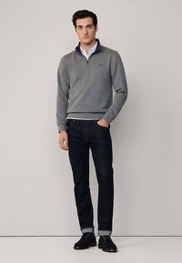 Maglione grigio con zip e colletto blu navy, camicia bianca sotto, jeans scuri e scarpe nere. Modello in piedi su sfondo neutro.