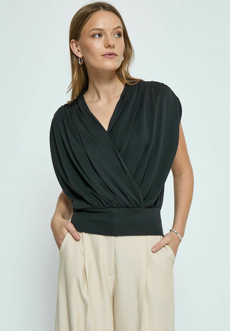 Top nero in stile avvolgente con tessuto plissettato e vita a costine, abbinato a pantaloni chiari beige a vita alta.