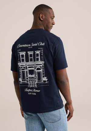 Férfi, aki sötétkék pólót visel, melyen fehér vonalas rajz látható egy épületről, kültéri ülőhelyekkel, valamint a "Downtown Social Club, Bedford Avenue, New York" felirat.