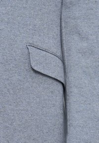 Grijs visgraatstof met een subtiele textuur, met een stijlvolle flapzak aan de linkerkant, wat functioneel detail aan het ontwerp toevoegt.