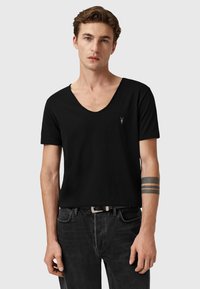 Camiseta negra de cuello en V hecha de una tela suave, de manga corta, con un pequeño logotipo bordado en el pecho, combinada con jeans oscuros.