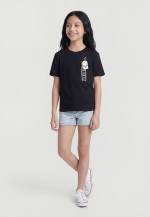 Νεαρή κοπέλα όρθια και χαμογελαστή, φοράει μαύρο γραφικό μπλουζάκι με ετικέτες, ανοιχτόχρωμα τζιν σορτς και λευκά αθλητικά παπούτσια μπροστά σε ένα απλό φόντο.