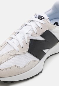 White athletic shoe featuring a combination of mesh and suede materials. The design includes a prominent black logo and textured accents.