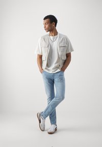 Light beige vest over a white t-shirt, light blue jeans, and white sneakers with black and brown accents. Simple, casual design.