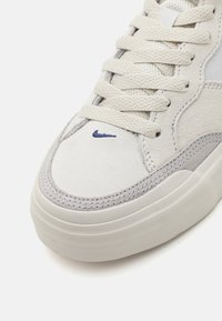 Hvit semsket sneaker med teksturert overflate, grå detaljer og en liten blå logo nær tåen. Har flate lisser og en tykk såle.