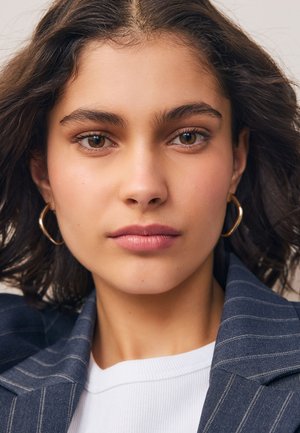 Close-up of a face with medium-length wavy dark hair, wearing gold hoop earrings and a navy pinstriped blazer over a white shirt.