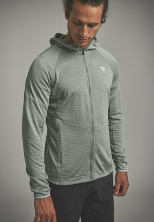 Mann trägt eine hellgraue Kapuzenjacke mit Reißverschluss und schwarze Hose, schaut nach unten und steht vor einem einfarbigen Hintergrund.