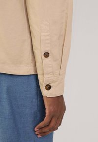 Beige Hemd mit Knopfverschluss und spitzen Manschetten, akzentuiert durch zwei dunkle Knöpfe. Der Stoff hat eine glatte Textur und wird mit blauen, strukturierten Hosen kombiniert.