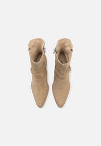Bottines en daim beige avec un design à bout pointu, dotées d'une fermeture éclair sur le côté et d'une sangle décorative autour de la tige supérieure.