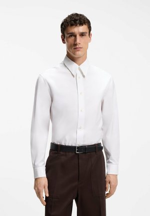 Mężczyzna ubrany w dopasowaną białą koszulę na guziki, włożoną do ciemnobrązowych spodni z wysokim stanem i czarnym paskiem, stojący na jednolitym tle.