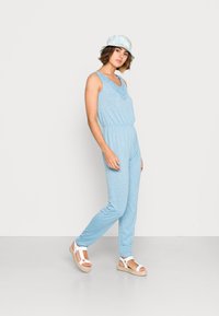 Macacão sem mangas de cor azul claro com detalhes em renda, cintura elástica e pernas ajustadas, combinado com um chapéu branco em estilo balde e sandálias.