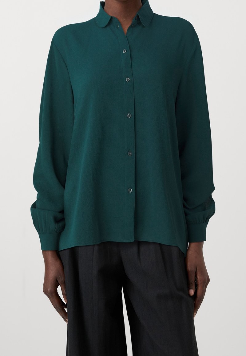 Blouse à manches longues vert foncé boutonnée avec col, portée avec un pantalon noir ample sur une personne devant un fond uni.