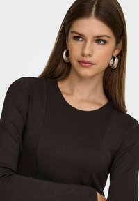 Top marrone a maniche lunghe con scollo rotondo, caratterizzato da un tessuto testurizzato e cuciture sottili. Modella che indossa orecchini a cerchio trasparenti.