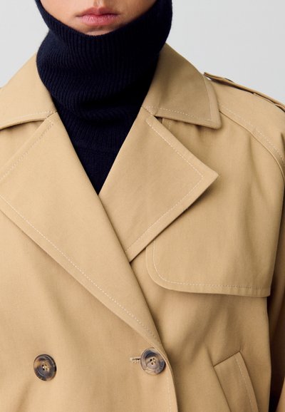 Trench-coat double boutonnage tan avec grands revers, couture crème et deux boutons marron. Superposé sur un col roulé côtelé bleu marine.