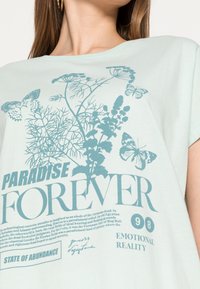 Mentolové bavlněné tričko s grafickým potiskem botanických prvků a motýlů v tmavě modré. Volnější střih s krátkými rukávy.