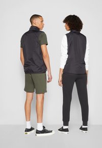 To personer iført svarte ermeløse vester med en glatt tekstur. Den ene har på seg olivengrønne shorts, mens den andre har svarte bukser. Begge har på seg uformelle joggesko.