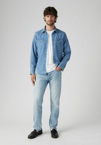 Camisa de mezclilla en azul claro, de manga larga, con botones a presión, y dos bolsillos en el pecho. Combinada con jeans azul claro y zapatos negros.