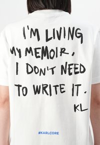 En person som har på seg en hvit T-skjorte med svart tekst på ryggen som sier "JEG LEVER MIN MEMOAR, JEG TRENGER IKKE Å SKRIVE DET NED." og blå hashtag #KARLCORE.