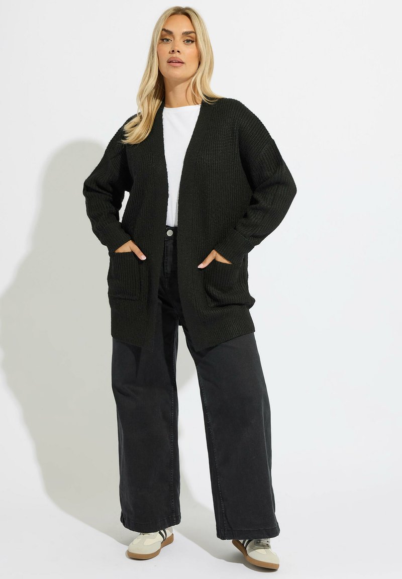 Svart stickad cardigan med öppen front, ribbad textur, stora fickor, bärs över en vit skjorta och matchas med vida svarta byxor.