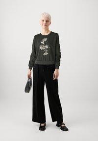 Sötétzöld pulóver grafikus nyomattal, fekete, bő szárú nadrággal és fekete cipővel párosítva. Kiegészítők közé tartozik egy kis, textúrázott táska.