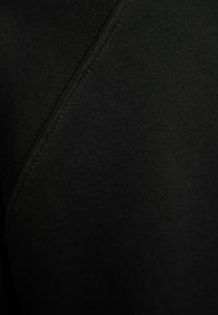 Detail černé látky s hladkou texturou a viditelnými stehy, s jemným leskem. Design má čistý, jednoduchý střih.