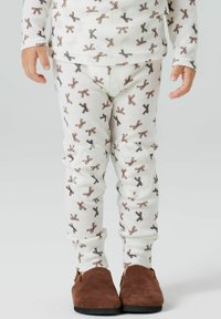 Ensemble de pyjama blanc avec un motif de nœud marron répété. Tissu doux, design ajusté, manches longues et poignets élastiques. Chaussures en daim marron.