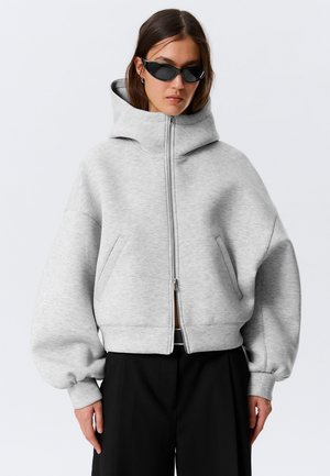 Kvinde iført oversized lysegrå lynlåshættejakke med høj krave, sorte solbriller og løstsiddende sorte bukser, stående mod en ensfarvet baggrund.