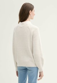 Cremfarbener, gerippter Pullover mit hohem Kragen, überschnittenen Schultern und langen Ärmeln. Kombiniert mit hellblauen Jeans für einen lässigen Look.