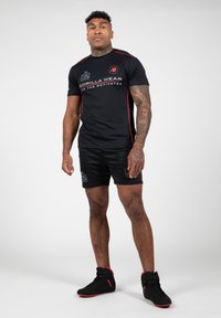 T-shirt de sport noir avec des accents rouges et un logo imprimé, associé à un short noir avec un logo. Porté avec des chaussures noires.
