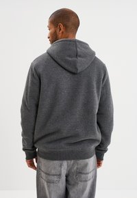 Sudadera gris con capucha de una sola capa, puños y dobladillo acanalados, y una textura suave. Lleva puestas unas jeans de mezclilla gris claro.
