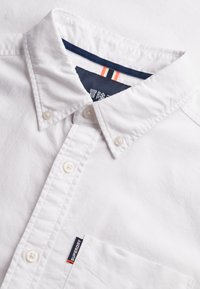 Chemise blanche boutonnée avec boutons sur le col, poche poitrine et étiquette marine portant l'inscription « Superdry ». Détail intérieur du col rayé bleu marine et orange visible.