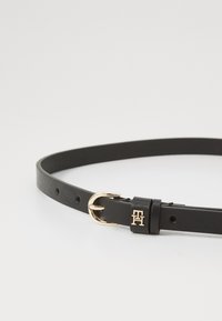 Cintura in pelle nera con fibbia in metallo dorato, dotata di numerosi fori di regolazione e un accento del logo vicino alla fibbia.