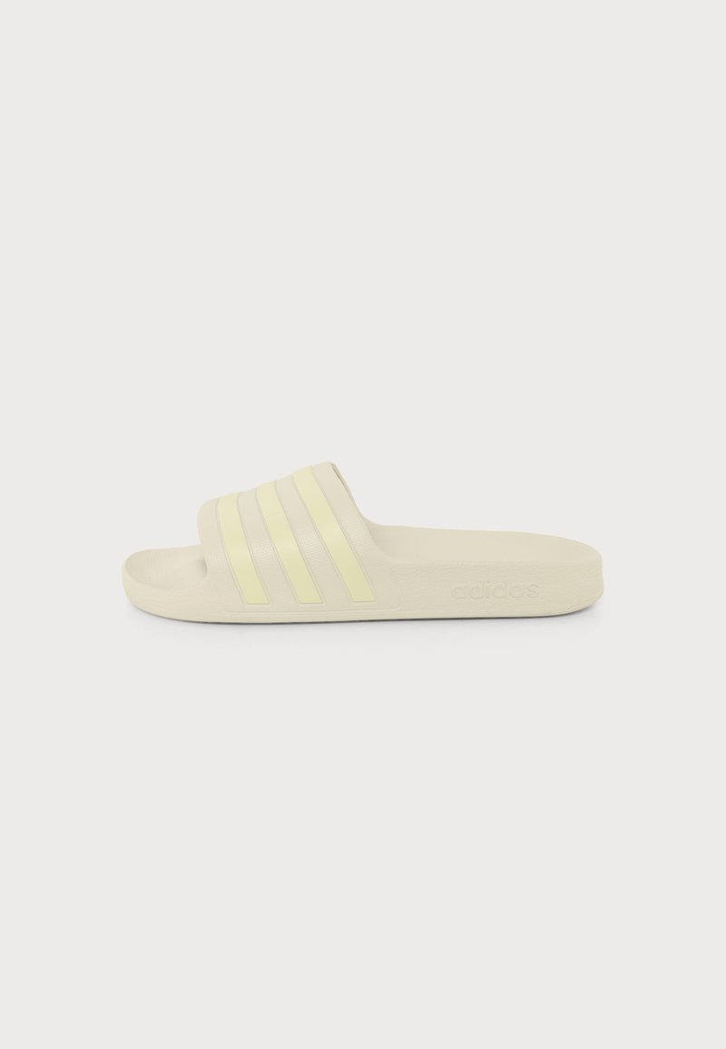 Ένα μπεζ Adidas σαγιονάρι με τρεις ανοιχτό κίτρινες ρίγες στο λουράκι, εμφανίζεται από το πλάι σε λευκό φόντο.