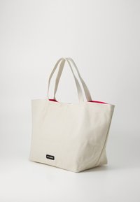Stofftasche in cremfarbener Ausführung mit kurzen Tragegriffen, ausgestattet mit einem lebhaften roten Innenfutter und einem kleinen schwarzen Logoetikett auf der Vorderseite.