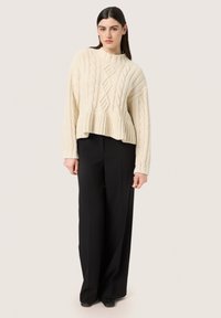 Creme kabelstrik sweater med rund halsudskæring og ribbet kant, parret med højttaljede sorte bukser med brede ben og sorte sko.