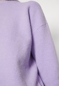 Gros plan sur une personne portant un pull lavande doux et texturé avec un col montant et des manches longues.