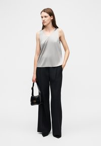 Mujer con blusa sin mangas plateada y pantalones negros de pierna ancha, sosteniendo un bolso negro, de pie sobre un fondo blanco liso.