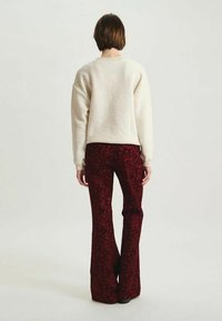 Une personne avec les cheveux bruns courts se tient de dos, portant un pull crème et un pantalon évasé à motifs rouge foncé sur un fond clair.