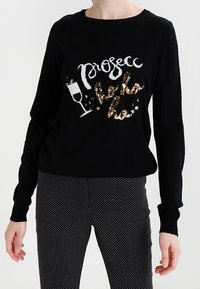 Černý svetr s dlouhým rukávem s nápisem "Prosecco hello ho" a motivem skleničky na šampaňské, spojený s černobílými puntíkovanými kalhotami.