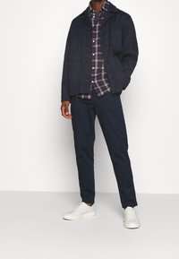 Giacca blu navy con chiusura a bottoni, indossata sopra una camicia a quadri, abbinata a pantaloni scuri e sneakers bianche. Tessuto morbido e fitting strutturato.