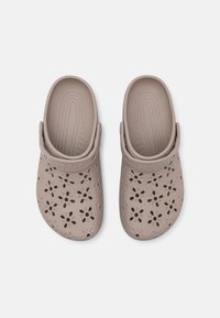 Kerged pruunid puukotid lõigatud lillemotiividega, valmistatud paindlikust materjalist. Omab taga rihma ja tekstureeritud jalapinda mugavuse jaoks.