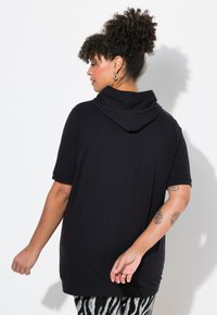 Felpa nera con cappuccio, maniche corte, vestibilità rilassata, texture liscia e cuciture sottili. Indossata con pantaloni a fantasia con strisce di zebra.