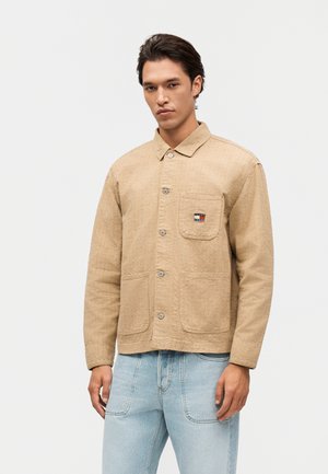 Man draagt een beige getextureerd jack met knopen en drie voorzakken, gecombineerd met lichtblauwe spijkerbroek, staand tegen een effen achtergrond.