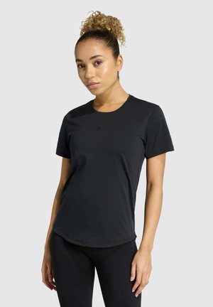 Donna con capelli ricci raccolti in un alto chignon, che indossa una maglietta aderente nera a maniche corte e pantaloni neri, in piedi davanti a uno sfondo chiaro uniforme.